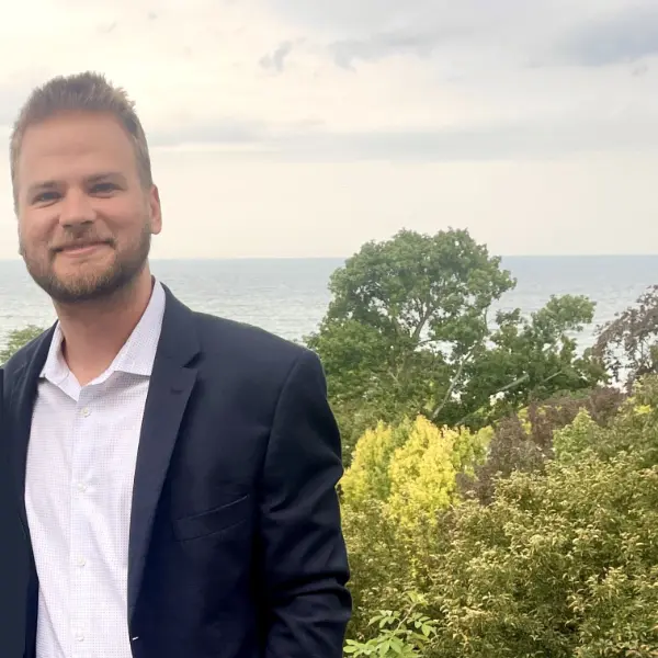 Man with short blonde hair smiling earing a dark blue blazer and white button up shirt outside with the a lake in the background